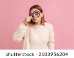 Portrait of funny blond woman standing, holding magnifying glass and looking at camera with big zoom eye, verifying authenticity, wearing white sweater. Indoor studio shot isolated on pink background.