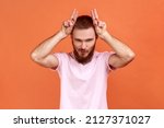 Small photo of Portrait of angry bully bearded man showing bull horn gesture with fingers over head, looking hostile and threatening, wearing pink T-shirt. Indoor studio shot isolated on orange background.