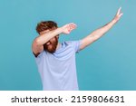 Small photo of Portrait of successful young bearded man showing dab dance pose, famous internet meme of triumph, performing dabbing trends with hand gesture. Indoor studio shot isolated on blue background.