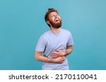 Portrait of positive overjoyed bearded man laughing happily at something keeps hands on belly, smiles broadly, expressing positive emotions. Indoor studio shot isolated on blue background.