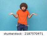 Portrait of man with Afro hairstyle wearing orange T-shirt showing empty pockets and looking frustrated about loans and debts, has no money, jobless. Indoor studio shot isolated on blue background.