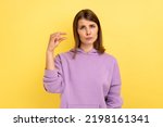 Small amount. Portrait of adorable beautiful woman showing little bit gesture with hand and looking pleadingly, wearing purple hoodie. Indoor studio shot isolated on yellow background.
