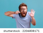 Small photo of Portrait of funny bearded man holding magnifying glass and looking at camera with big zoom eye, curious face, waving hand, saying hi. Indoor studio shot isolated on blue background.