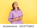 Portrait of happy woman with dark hair holding belly, laughing, hearing funny joke or anecdote, being in good mood, wearing purple hoodie. Indoor studio shot isolated on yellow background.