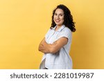 Portrait of satisfied delighted happy woman with dark wavy hair standing with folded hands and looking at camera with toothy smile. Indoor studio shot isolated on yellow background.