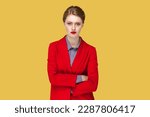 Portrait of serious strict woman with red lips standing standing with crossed arms, looking at camera, having bossy expression, wearing red jacket. Indoor studio shot isolated on yellow background.