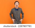 Small photo of Portrait of shocked funny man nerd standing looking up with big eyes and widely ope mouth, wearing shirt with blue bow tie and white glasses. Indoor studio shot isolated on orange background.