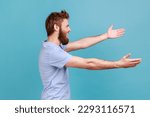 Small photo of Free hugs, come into my arms. Side view of happy bearded man stretching hands to camera and smiling broadly, going to embrace, share love. Indoor studio shot isolated on blue background.