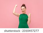 Portrait of extremely happy positive woman standing with raised clenched fists, winning lottery, celebrating her success, wearing green dress. Indoor studio shot isolated on pink background.