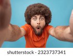 Portrait of astonished man with Afro hairstyle wearing orange T-shirt looking with open mouth and big eyes, taking selfie, point of view photo, POV. Indoor studio shot isolated on blue background.