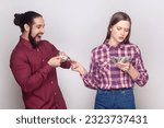Small photo of Portrait of serious rich confident woman and man gigolo standing together, wife giving money to her husband with indifferent face. Indoor studio shot isolated on gray background.