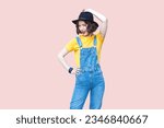 Portrait of funny positive confident hipster woman in blue denim overalls, yellow T-shirt and black hat, smiling to camera. Indoor studio shot isolated on light pink background.