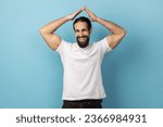 Small photo of Safety and house insurance. Portrait of man with beard wearing white T-shirt making roof gesture over head and smiling to camera with amiable expression. Indoor studio shot isolated on blue background