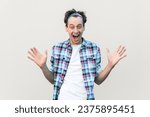 Small photo of Surprised attractive man keeps jaw dropped and eyes widely opened, has rapt attention, raised his arms, wearing blue checkered shirt and headband. Indoor studio shot isolated on gray background.