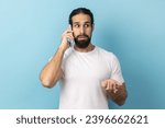 Small photo of Portrait of confused man with beard wearing white T-shirt standing talking on mobile phone, looking away with puzzlement, hearing breaking news. Indoor studio shot isolated on blue background.