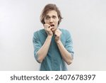 Small photo of Man feels anxious and surprised, bites finger nails in puzzlement, looks nervously at camera, worries before presenting project, wearing blue shirt. Indoor studio shot isolated on gray background.