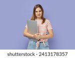 Portrait of cheerful happy optimistic blond woman wearing striped T-shirt standing with closed laptop, finishing her work, being satisfied. Indoor studio shot isolated on purple background.