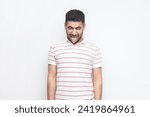 Small photo of Portrait of foolish crazy childish bearded man wearing striped t-shirt standing with foolish facial expression, sticking tongue out. Indoor studio shot isolated on gray background.