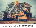 Small photo of Caucasian Male Contractor Kneeling by Toolboxes Just Taken Out of His Pickup Truck Preparing the Necessary Tools for the Full Day of Work at the Building Site. Industrial Theme.