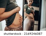 Small photo of Unhappy young woman standing in front of a mirror and holding hands on her bloating stomach.
