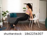 Small photo of Attractive young woman exercising in her home. Sporty woman in her 20s doing tricep dips using a chair in the living room