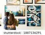 Beautiful couple seen from behind is hugging while admiring the paintings and colorful canvases hanging on the wall of the gallery 
