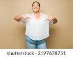 Confident hispanic plus size woman smiling pointing down to copy space ad against a pastel yellow background