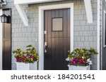 Angled view of glossy black front door to a family home; The door is made of vertical wood boards, with a window. It is framed by two flower planters, gray shingles of the house, and a door mat. 