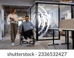 Young business colleagues, collaborative business colleagues, including a person in a wheelchair, walk past a modern glass office corridor, illustrating diversity, teamwork and empowerment in the