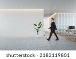 Small photo of Side view of young businessman walking in modern concrete and wooden coworking office interior with empty mock up place on wall, furniture and equipment. CEO and executive concept