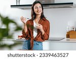 Shot of beautiful happy woman eating a yoghurt while looking forwards in the kitchen at home