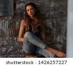 Elegant casual dressed woman sitting at high chair against brick wall. Girl in jeans and black top with long curly hair. Loft interior. Evening sun light. Full length portrait