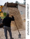 Small photo of a bulldozer pours macadam from a bucket onto the ground at a construction site in front of gray-haired worker with shovel.