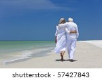 Rear view of a senior man and woman couple walking arms around each other on a deserted tropical beach with bright clear blue sky, s3niorlife