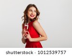 Portrait of a cheerful young woman dressed in red dress holding glass of champagne and looking away isolated over white background