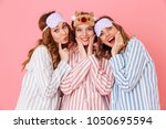 Three beautiful young girls 20s wearing colorful striped pyjamas and sleeping masks having fun during girlish sleepover isolated over pink background