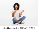 Portrait of smiling young afro american woman pointing fingers away while sitting on a floor with legs crossed isolated over white background