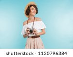 Portrait of a smiling young woman in summer hat standing with photo camera and looking away at copy space isolated over blue background