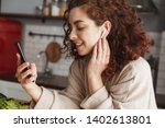Portrait of cute european woman with earpods listening to music on cellphone while cooking in kitchen interior at home