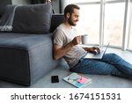 Attractive happy smart young man sitting on a floor in the living room, working on ;laptop computer, drinking coffee