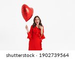 Excited beautiful girl wearing red dress posing with heart balloon isolated over white background