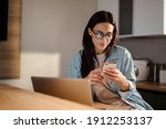 Serious charming woman using smartphone while working with laptop at home