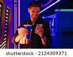 Small photo of Young bristle man using cellphone while standing with soda and popcorn in cinema