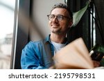 Portrait of adult handsome smiling man in glasses looking out the window while sitting with book in cafe