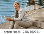 Asian mature woman in tracksuit using cellphone and sitting on stairs during workout outdoors