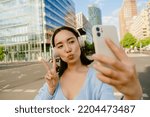 Small photo of Young beautiful asian girl with puckered lips taking selfie on phone and showing victory gesture, while standing in the city on the street