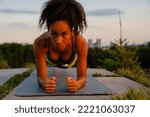 Small photo of Portrait of young beautiful focused curly african woman with puckered lips holding plank position and looking aside, on yoga mat outdoors during sunset