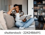 Young positive smiling african man wearing plaid shirt reading book while sitting on couch at home