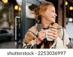 Joyful young blonde woman in warm plaid drinking hot tea while stanging outdoors at city street