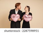 Young loving positive couple wearing black clothes holding heart-shaped balloons and kissing isolated over beige studio wall